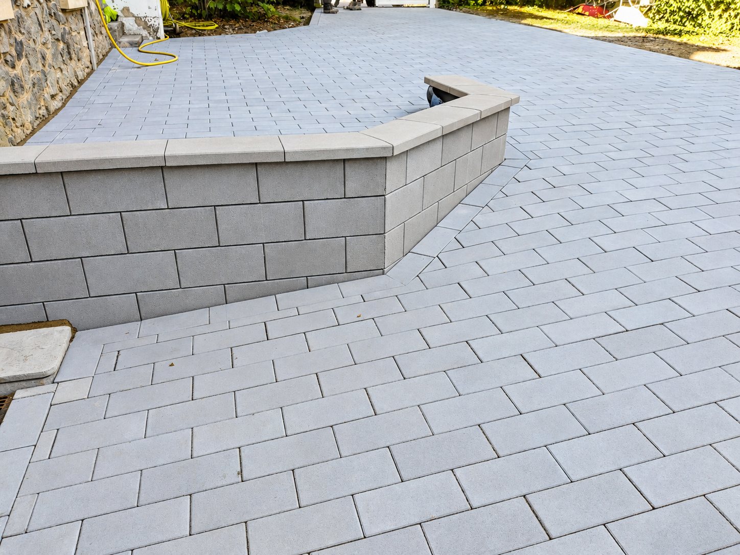 Une allée récemment pavée de grands pavés gris clair, bordée d'un muret en blocs de béton avec couvertine, le long d'un mur en pierre.