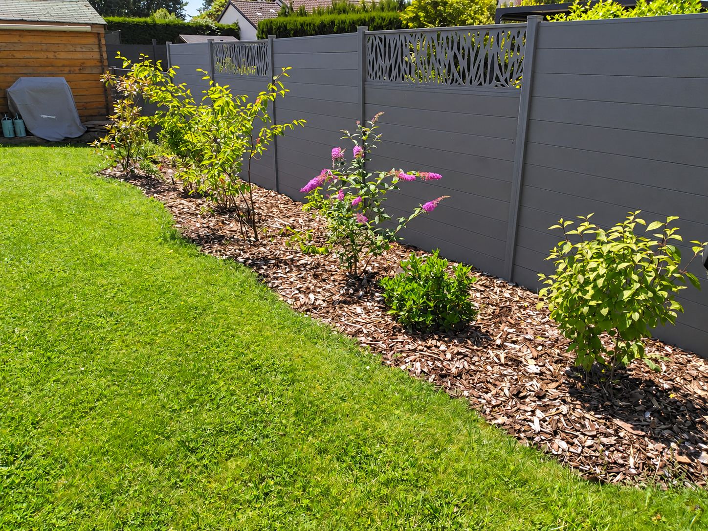 Une clôture de jardin grise en lames composites avec panneaux ajourés décoratifs, longée d'un massif de paillage où poussent des arbustes dont un buddleia en fleurs, au bord d'une pelouse.