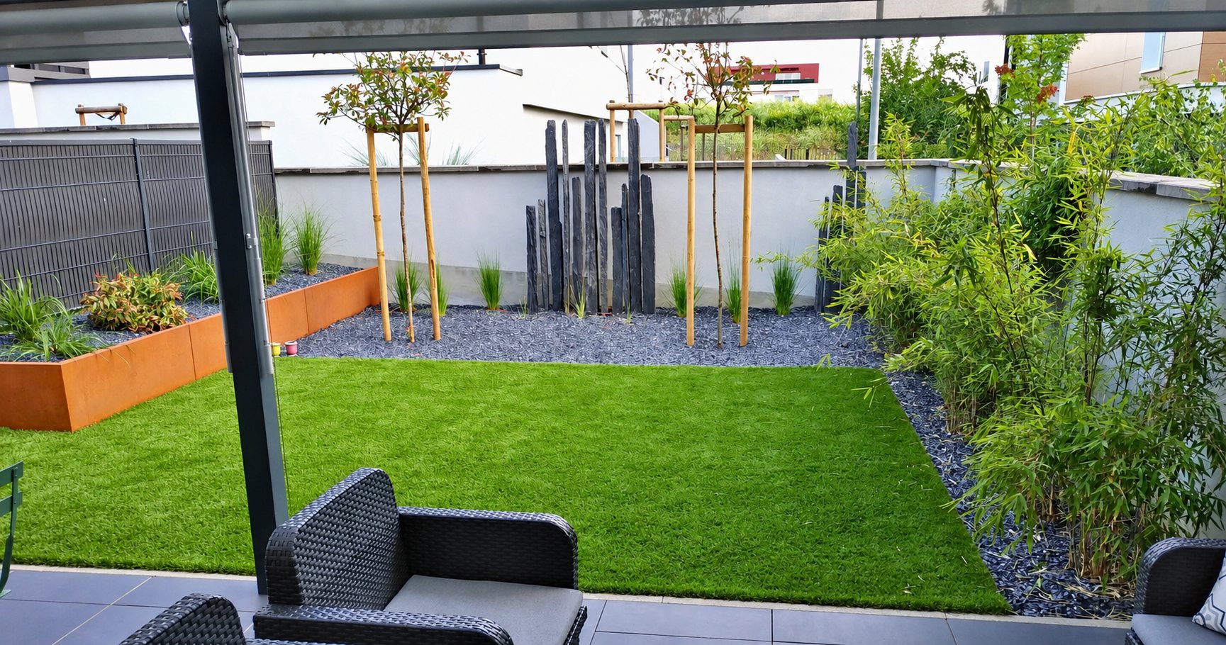 Petit jardin de ville aménagé avec une pelouse synthétique, des lames d'ardoise, des bambous et une jardinière en acier corten, vu depuis une terrasse avec fauteuils en résine tressée.