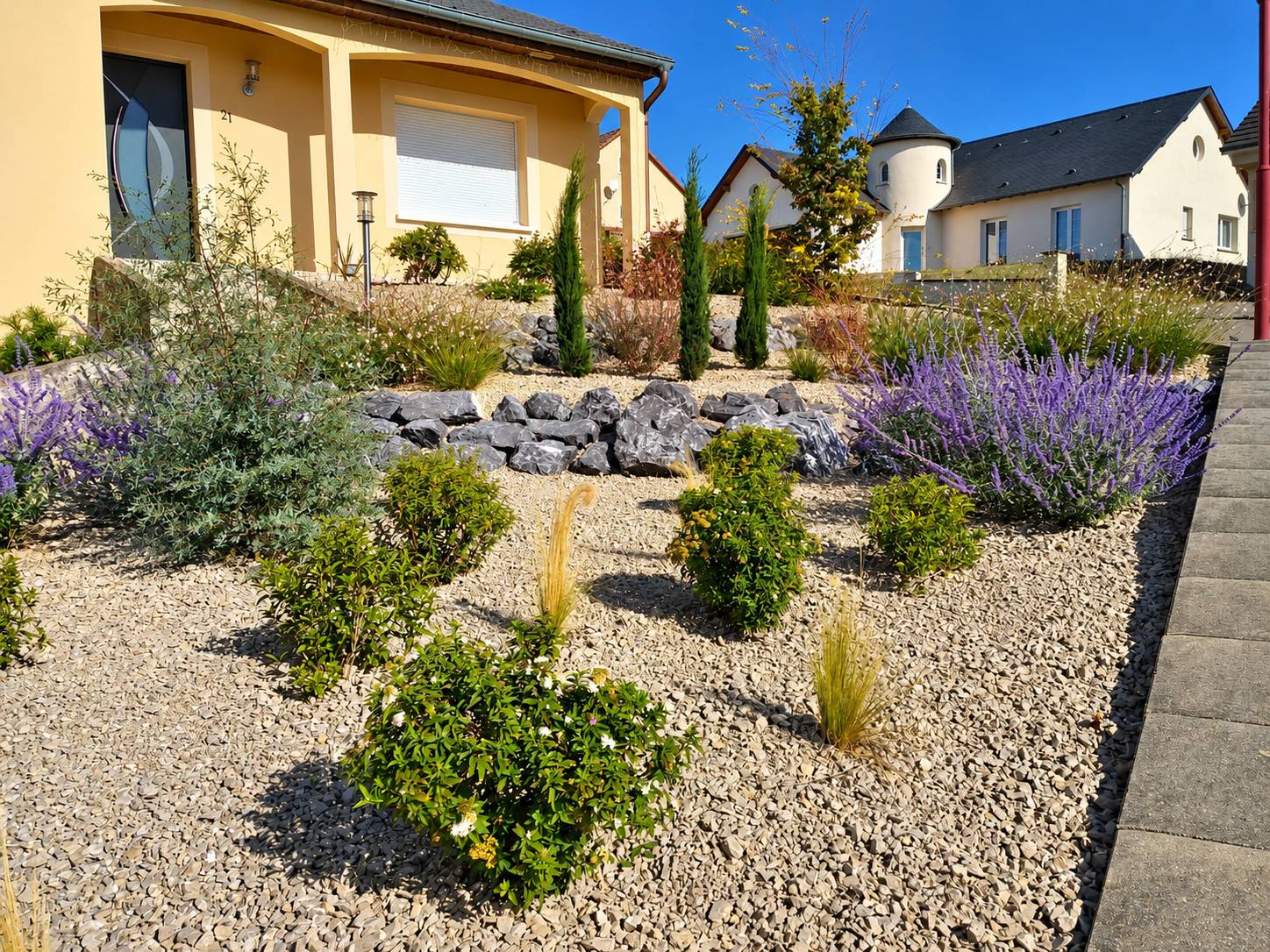Aménagement paysager d'un jardin de devant en gravier avec rochers, cyprès, lavande et arbustes, devant une maison jaune.