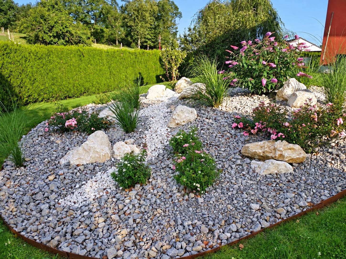 Massif de jardin aménagé avec des galets gris, un chemin de pierres blanches, de gros rochers, des graminées et des buddleias roses, bordé d'une haie.