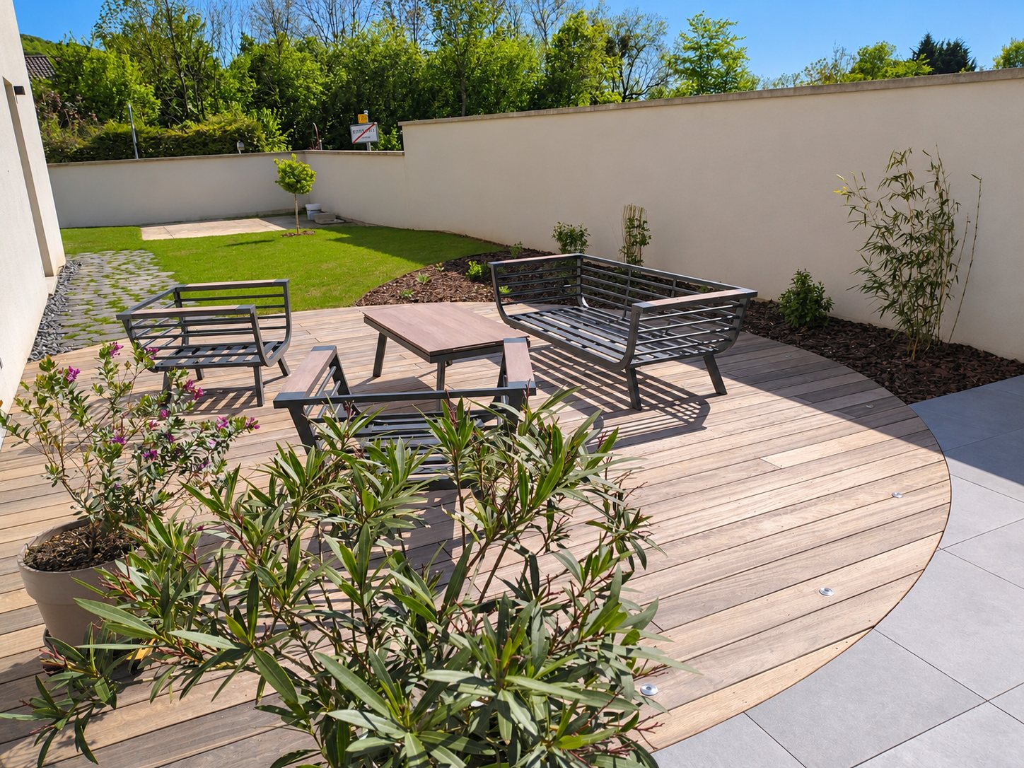 Une terrasse en bois de forme arrondie meublée d'un salon de jardin métallique, entourée de massifs, de pelouse et d'un haut mur crème.